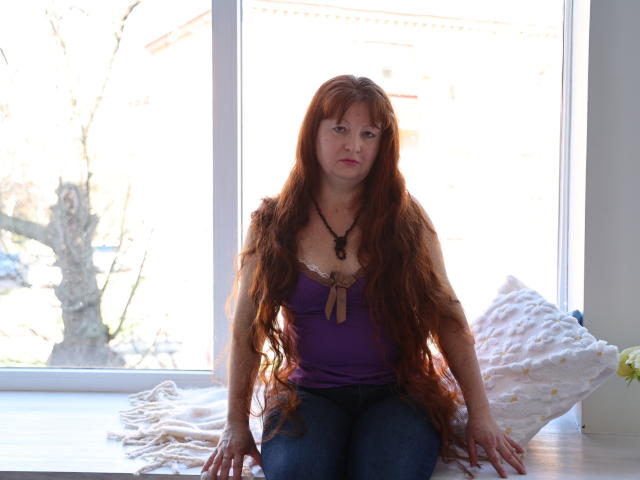 OVanessaHotly with long red hair, purple top, and blue jeans, sitting on a white surface, looking serious, wearing a necklace, hands on her knees, natural light.