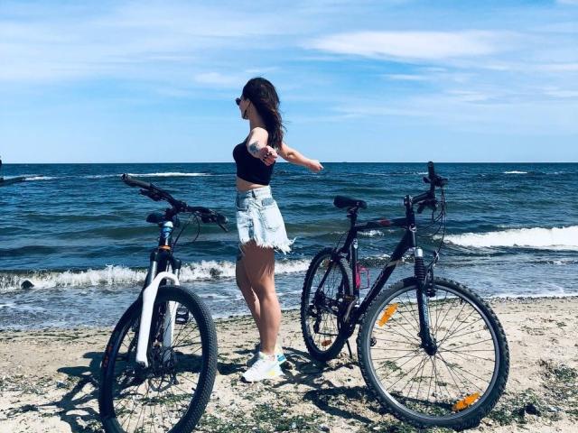 Milagres with medium tits, wearing a black crop top and frayed denim shorts, stands by two mountain bikes on a sandy beach with a blue ocean in the background.