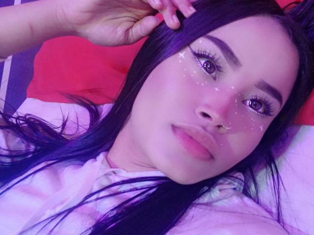 KarolPalacios has purple hair, a nose ring, glitter on her face, and is lying down with a hand in her hair, looking at the camera with a subtle smile.