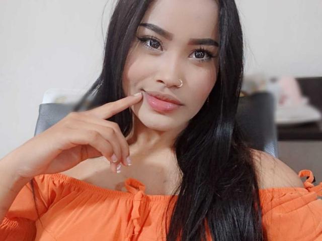 KarolPalacios with long black hair, wearing an off-shoulder orange top, has a nose ring and is touching her chin with her finger, looking slightly smug.