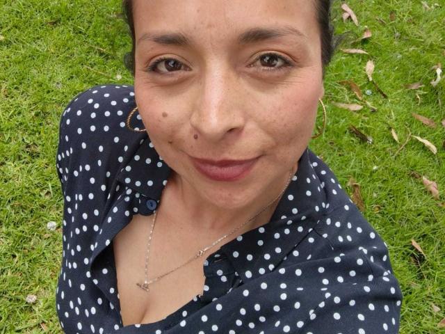 StangelarRivera with brown eyes, small nose, and subtle smile, wearing a navy polka dot shirt, necklace, and no makeup, sitting on green grass, showing a small beauty mark.