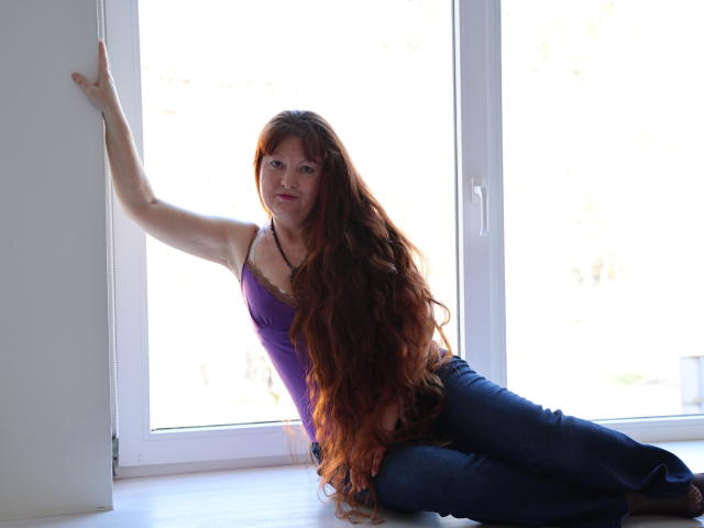 OVanessaHotly with long red hair, purple tank top, and blue jeans, leaning against a window, showing her tits, with a relaxed, sexy, and confident expression.