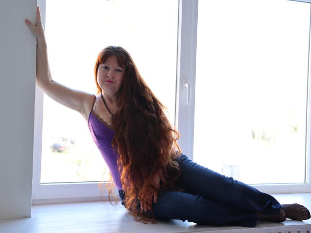 OVanessaHotly with long red hair, purple top, and blue jeans, sitting on window sill, arm raised, looking at camera, sunlight in background, sexy, slim, cute.