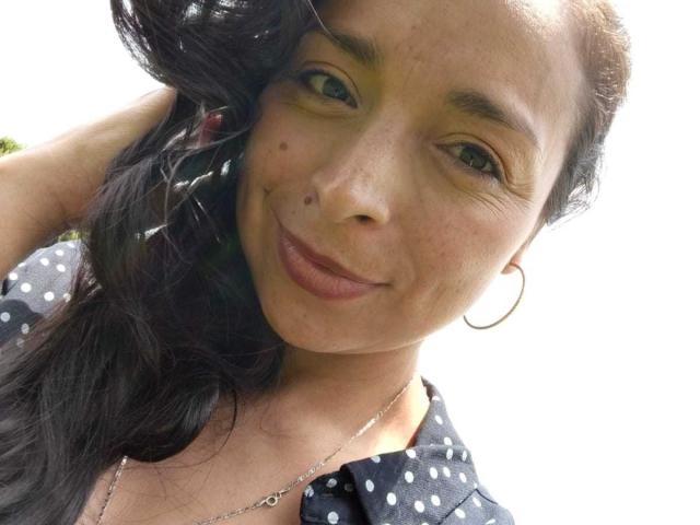 StangelarRivera with dark wavy hair, green eyes, and small moles on her cheek, wearing a polka dot shirt, hoop earrings, and a necklace, smiling slightly.
