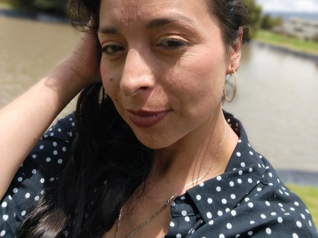 StangelarRivera has medium brown skin, dark hair, and small tits. She's wearing a black polka dot shirt, gold hoop earrings, and a necklace, with a slight, confident smirk.