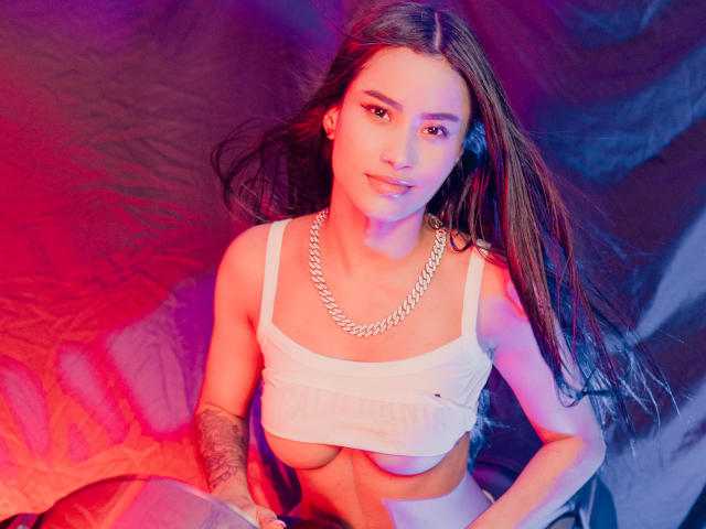 KarolPalacios with long brown hair, white crop top showing tits, chain necklace, tattooed arm, sitting, looking up, red and purple light.