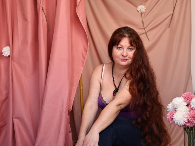 OVanessaHotly, with long red hair, pink lipstick, and a purple tank top, sitting on a chair, showing her tits, wearing a black bead necklace, in a pink and beige setting.