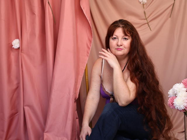 OVanessaHotly with long red hair, purple bra, and blue jeans, sitting, hand on chin, pink and beige curtains, white and pink flowers, looking slightly bored.