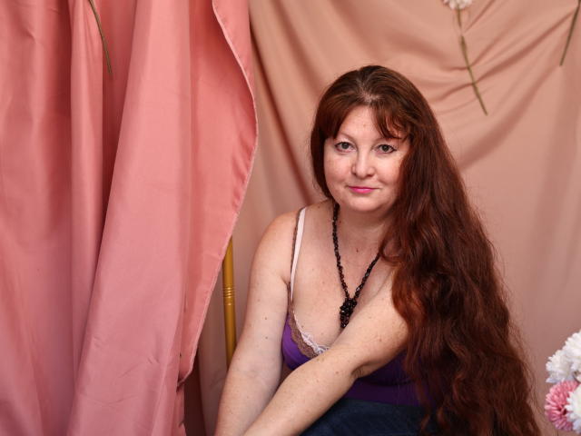 OVanessaHotly with long brown wavy hair, wearing a purple and white tank top, black bead necklace, and a slight smile, sitting in front of pink curtains.