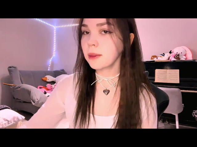 LezlieKennady with long brown hair, white choker, and heart pendant, wearing a white top, sitting in a room with blue string lights and stuffed animals.