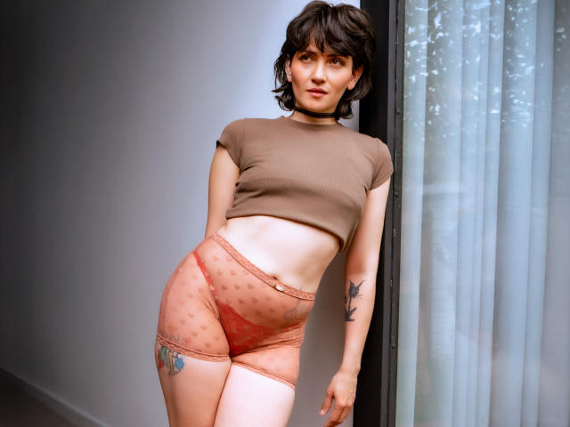 IvetteDurand has short brown hair, a choker, a brown crop top, and sheer pink polka-dot panties, showing a tattoo on her left thigh and a black tattoo on her right arm.