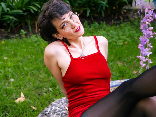 IvetteDurand with short black hair, glasses, red dress, and black tights, sitting on rocks, green grass, and purple flowers, looking up with a slight smile. Her tits are visible.