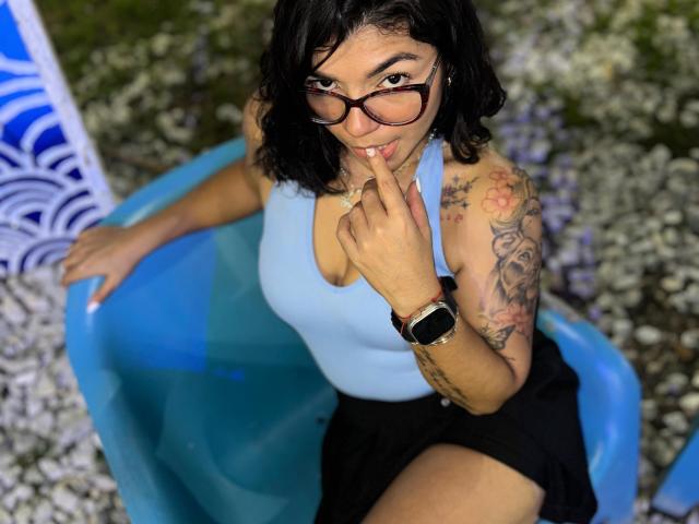 ValeryPierce, Latina, black wavy hair, glasses, blue halter top, black skirt, tattooed arm, finger to lips, blue plastic chair, black watch, sitting outdoors, looking up, smiling.