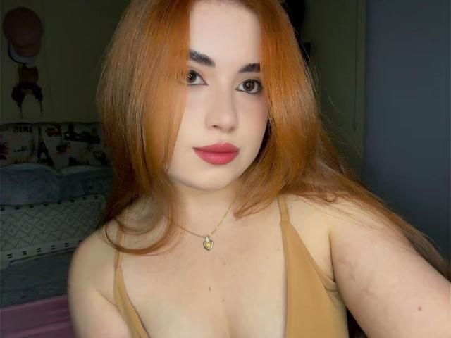 VicluX has long orange hair, pale skin, and red lipstick. She's wearing a tan top, showing her tits, and has a gold necklace. Her eyebrows are dark and thick.