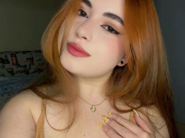 VicluX with orange hair, pink lipstick, and yellow nails, holding chest, wearing heart necklace, has black ear studs, and a slight smile. Boobs visible, head tilted.