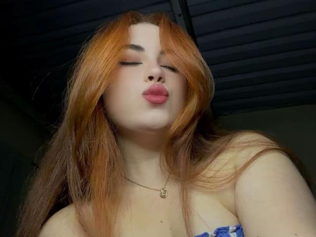 VicluX with long red hair, pouty pink lips, and a gold necklace, looking down at the camera with a slightly open mouth. Boobs visible, wearing a blue strap.
