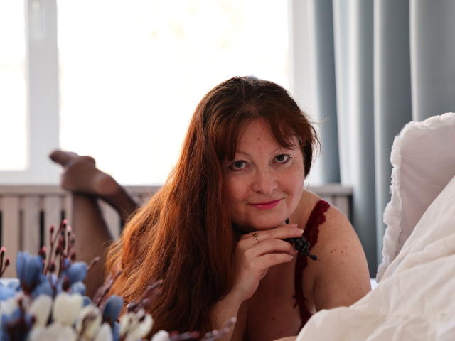 OVanessaHotly with red hair, lying on bed, smiling, hand under chin, wearing red lace bra, white pillow, blue and white flowers in foreground, legs up, black stockings.