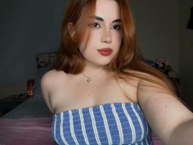 VicluX ha i capelli rossi, indossa un top a tubino a righe blu e bianche, che mette in mostra il suo seno abbondante, la pelle chiara e il rossetto rosso, con un leggero sorriso, in una stanza poco illuminata..