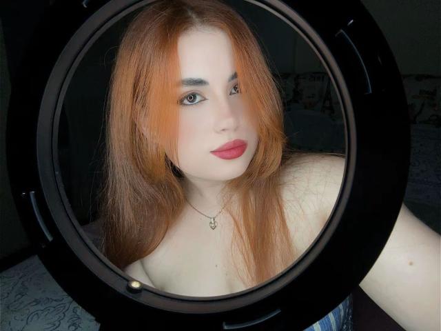 VicluX has red hair, pale skin, and red lipstick, looking through a ring light with black frame, showing off bare shoulders and a necklace with a small pendant.