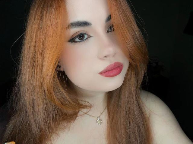 VicluX, con i capelli arancioni, la pelle chiara, l'eyeliner nero e il rossetto rosso, guarda leggermente di lato, indossa una collana a forma di cuore e mostra le spalle scoperte..