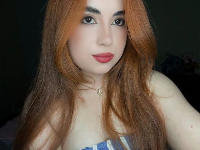 VicluX has long, red-orange hair, pale skin, and wears pink lipstick. She's looking at the camera with dark eyes, wearing a strapless top that shows her boobs.
