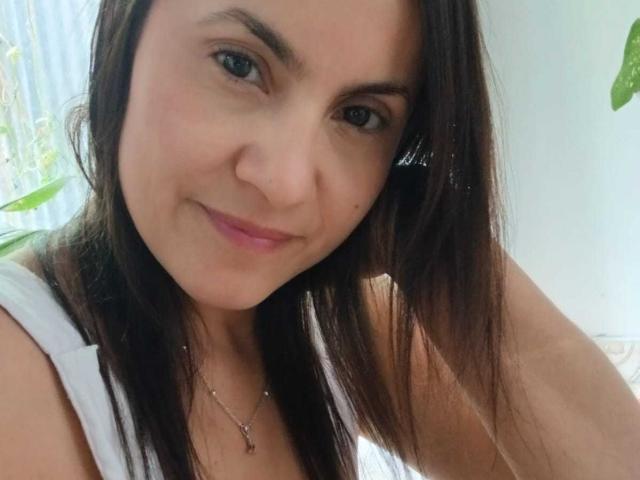SolaraDubois with brown hair, blue eyes, and pink lips, smiling slightly, wearing a white tank top and a necklace with a small pendant, in a bright room.