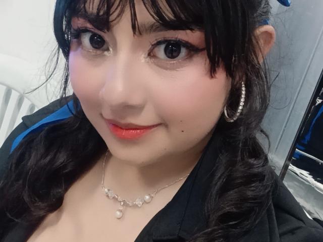 NekoPam has dark hair, big eyes with thick eyeliner, red lipstick, small pearl necklace, and silver hoop earrings. Her black top shows a hint of cleavage.
