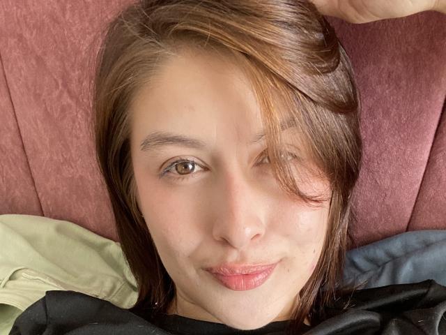 IsabellaPalacio with brown hair, winking, and puckering her lips, lying on a pink couch. Her face is centered, and she's wearing a black shirt.