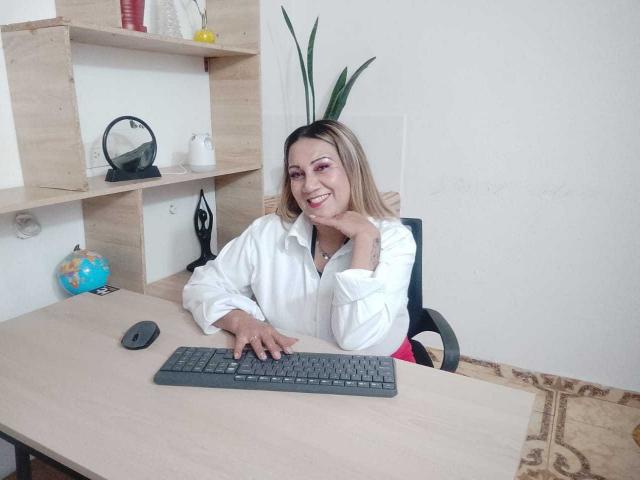 MatureYayita, with blonde hair, white shirt, and pink lipstick, sits at a desk, smiling, hand on cheek, fingers on keyboard, mouse beside her.