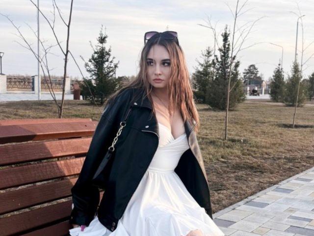 CarlotaLongNecker with long brown hair, wearing a black jacket over a white dress, sitting on a wooden bench, sunglasses on head, looking serious, grassy park background.