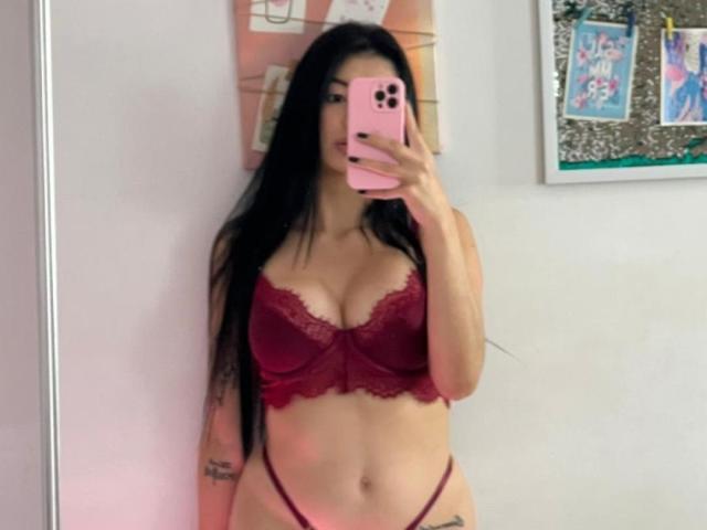 IsabellaJames with long black hair, wearing a red lace bra that shows her tits and a thong, holding a pink phone, standing in front of a mirror.