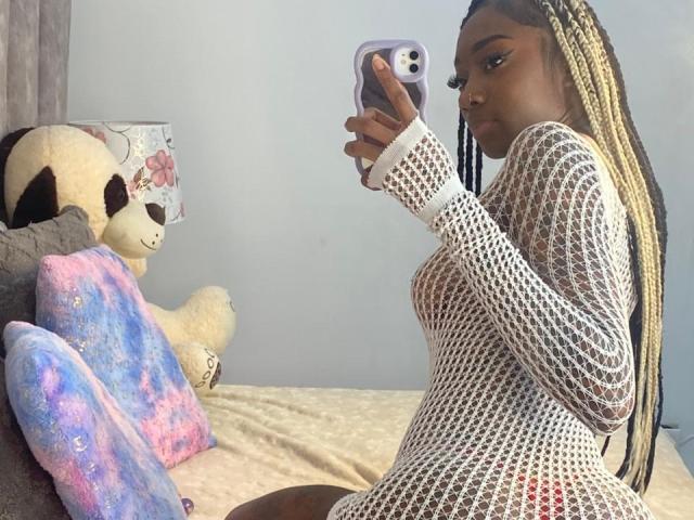 MilanCartier with dark skin, blonde braids, wearing a white mesh dress, showing her small tits and pussy outline, taking a mirror selfie, holding a purple phone.