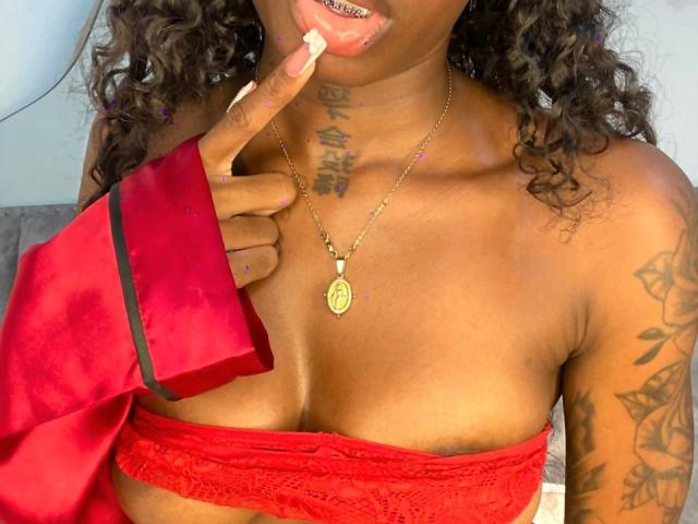 MilanCartier with curly hair, tongue out, red lace top, gold necklace, tattoos, finger on lips, red sleeve, dark skin, small boobs, gold teeth, nail art, seductive expression.
