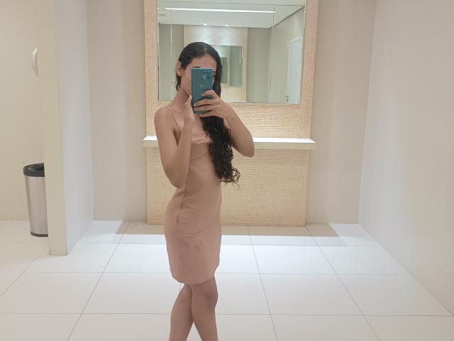 AngelRosa69 is naked, standing in a bathroom, taking a mirror selfie with her phone. She has long curly brown hair, covering her tits with her arm. Her ass is visible.