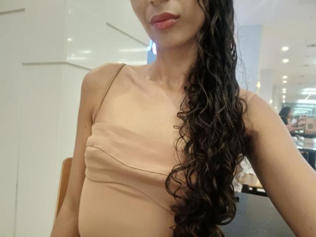 AngelRosa69 with long, wavy black hair, pink lipstick, and a tan, strapless top showing her small tits. Her skin is light, and she's standing indoors.