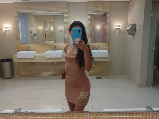 AngelRosa69 with long black hair, wearing a tight, tan dress, showing her ass, standing in a bathroom, taking a mirror selfie with a blue phone.