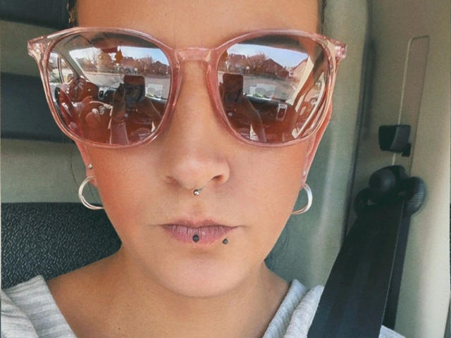 SpicyJenny - aantrekkelijk met roze getinte brillen, neusring, lipring en zilveren oorringen, draagt een veiligheidsgordel, reflectie in de brillen toont het interieur van de auto..