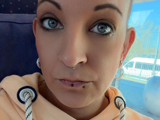 SpicyJenny-hot has blue eyes, a nose piercing, a lip piercing, and a small black lip stud. She's wearing a hoodie with white straps. Her face is slightly tilted.