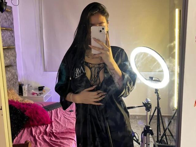 MaddyCarina, avec de longs cheveux noirs, portant une robe en satin noir qui révèle de la lingerie en dentelle, tenant un téléphone devant un miroir, avec un éclairage annulaire sur un trépied, et une main posée sur son ventre..