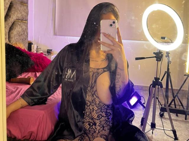 MaddyCarina with black lace bodysuit, large butt cutouts, black satin robe, holding phone, ring light, tattoo on right arm, in mirror selfie, pink bed in background.