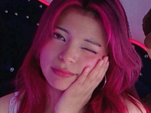 ShofiaWett with pink hair, winking, resting her hand on her cheek, smiling softly. She's wearing a white top and has pink highlights in her hair.