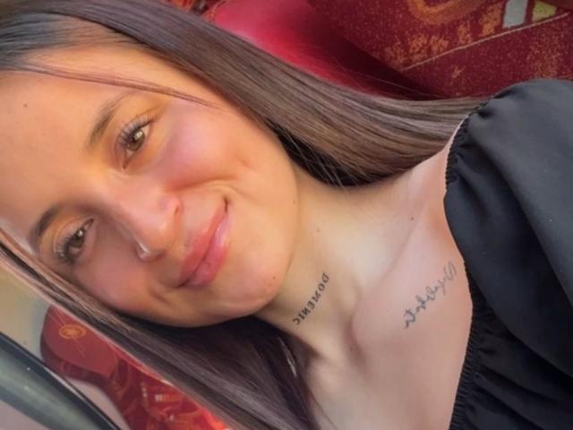 LucyArcy has a tattoo on her collarbone with cursive writing, brown hair, a black top, and a cute smile. Her tits are small and she's looking at the camera.