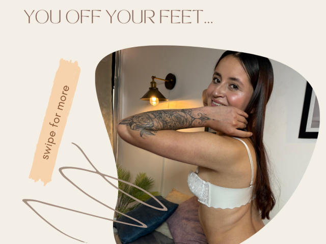 LucyArcy with tattooed arm, smiling, wearing white lace bra, showing off feet, brown hair, light skin, in cozy room, holding arm up, lamp in background.