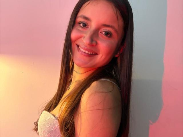 LucyArcy with long brown hair, smiling, wearing a white lace strap dress, has small tits, fair skin, and a subtle tattoo on her neck, in pink and orange lighting.