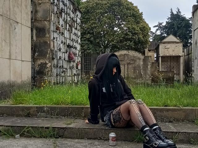 sopor66 sitting on concrete steps, black hoodie, face covered, long black hair, fishnet stockings, patterned shorts, black boots, can on ground, grass, trees, and old building in background.