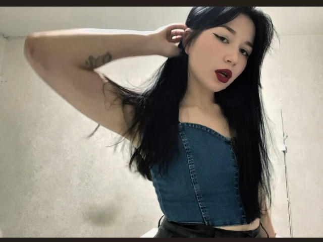 MertieDasinger with long black hair, wearing a blue denim strapless top, red lipstick, and black eyeliner, showing a tattoo on her right arm, hand in hair.