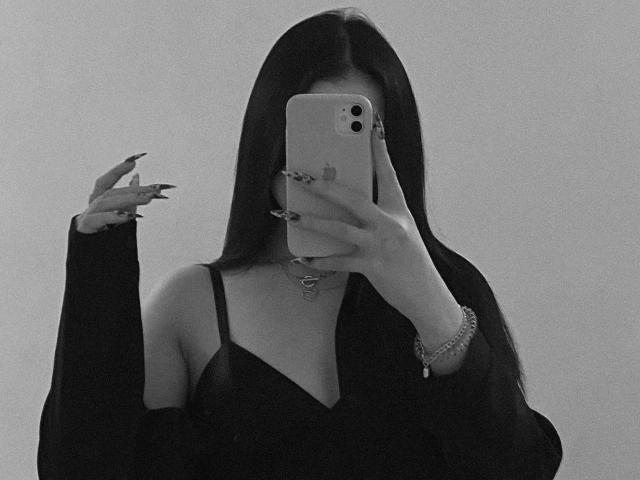 MertieDasinger with long black hair, wearing a black dress with spaghetti straps, taking a mirror selfie with a phone, nails long and pointed, choker necklace, bracelet on right wrist.