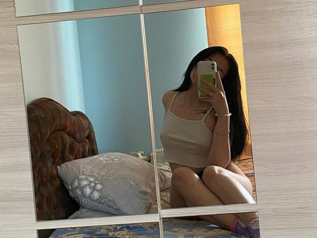 MertieDasinger with long black hair, white tank top, and no pants, sitting on a bed, taking a mirror selfie, showing her tits and pussy in the reflection.