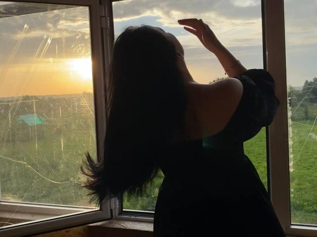 MertieDasinger with long dark hair, facing window, wearing black off-shoulder dress, raising arm, sun setting, glass reflecting light, green field outside, silhouetted against orange sky.