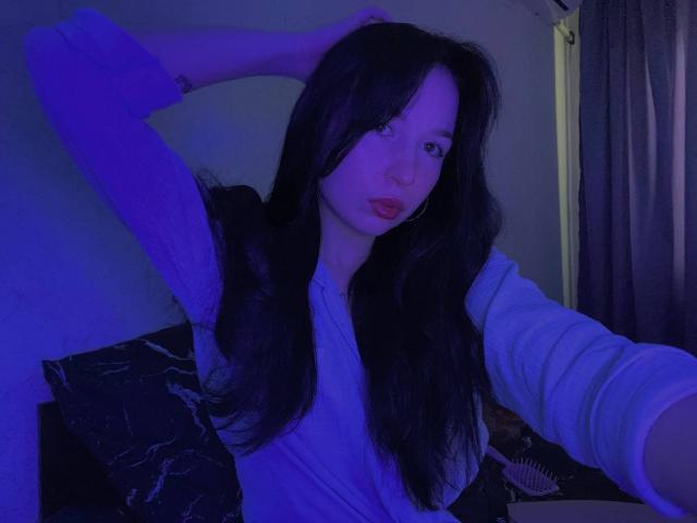 MertieDasinger with long black hair, blue-lit face, wearing a white long-sleeve shirt, hand in hair, slightly parted lips, looking at camera, dark curtains in background.